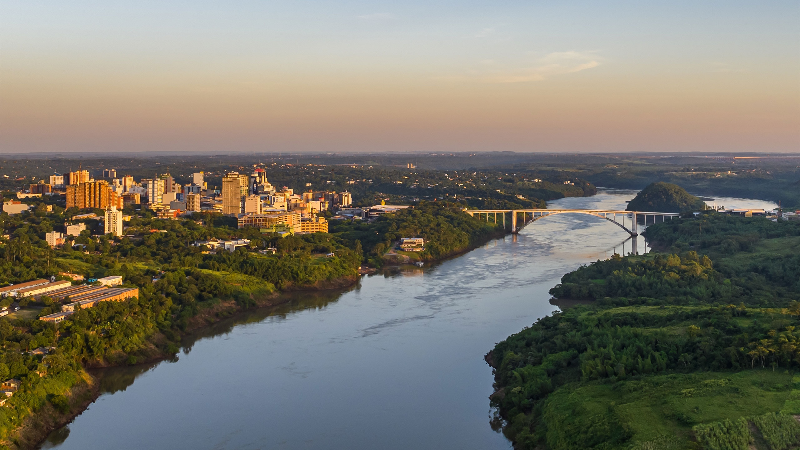 Vista aérea de um rio largo com um ponte de arco ligando duas margens verdes e uma cidade ao fundo durante o pôr do sol.
